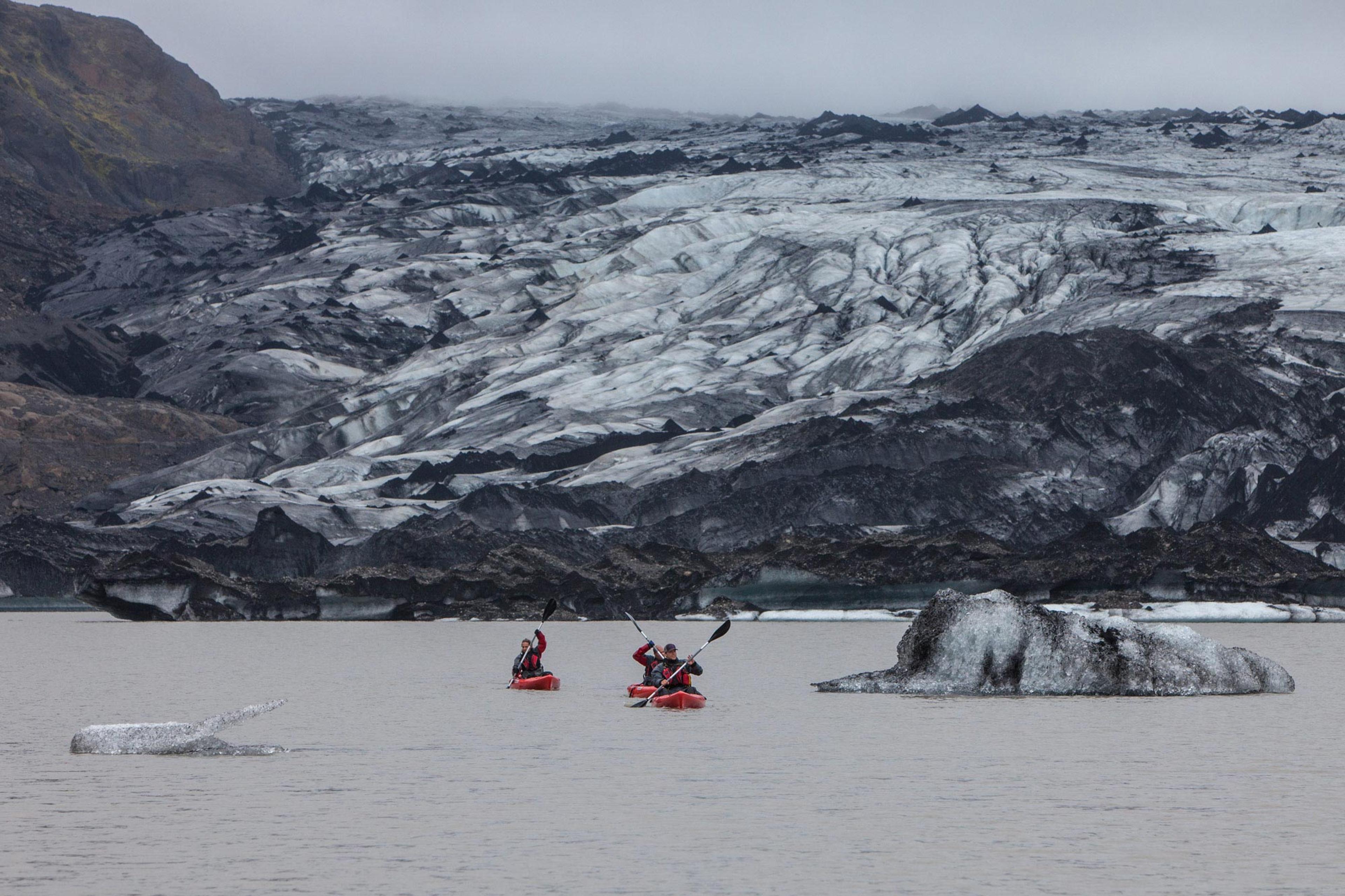 People in red kayaks paddlilng on a glacier lagoon with a stunning glacier in the background.