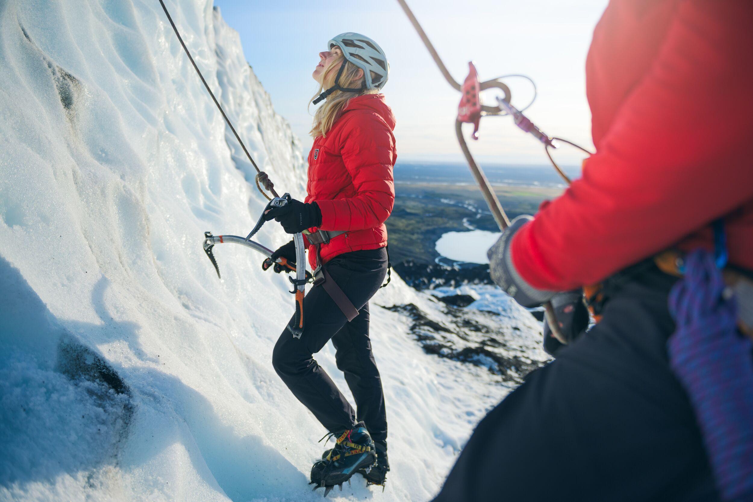 Two climbers equipped with helmets, harnesses, and ice axes preparing to ascend an icy slope on a glacier, with one climber looking up while roped in for safety.