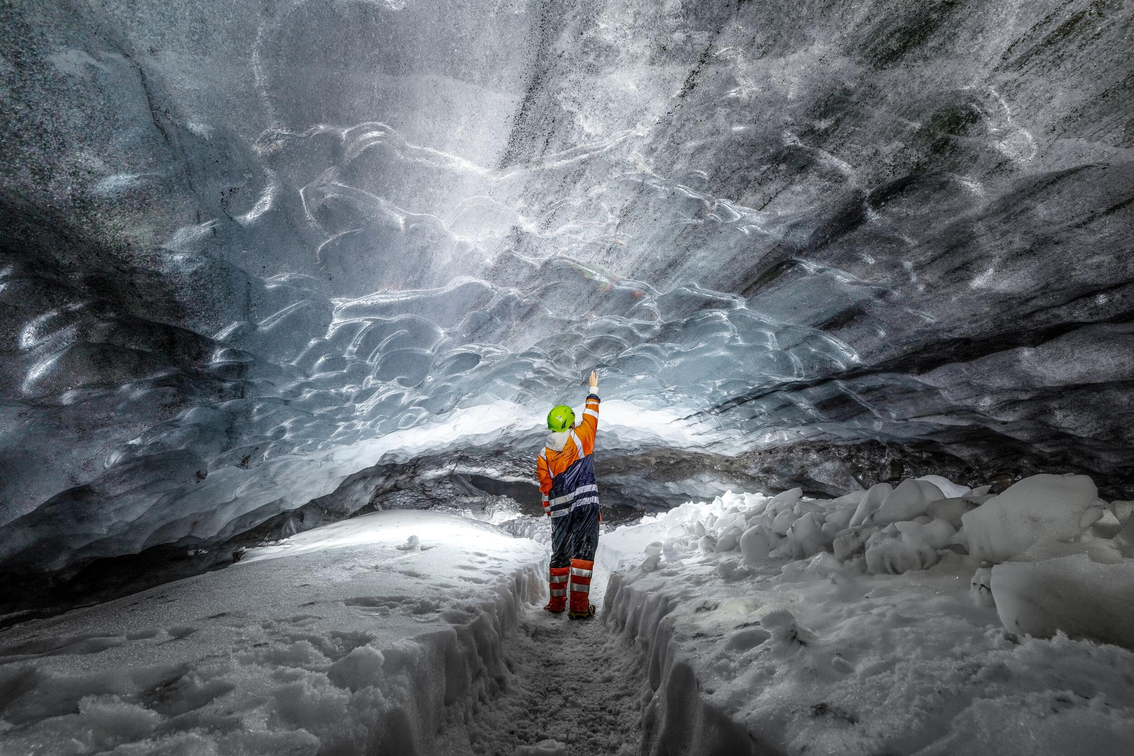 A person wearing protective gear reaching out to the ceiling inside an ice cave