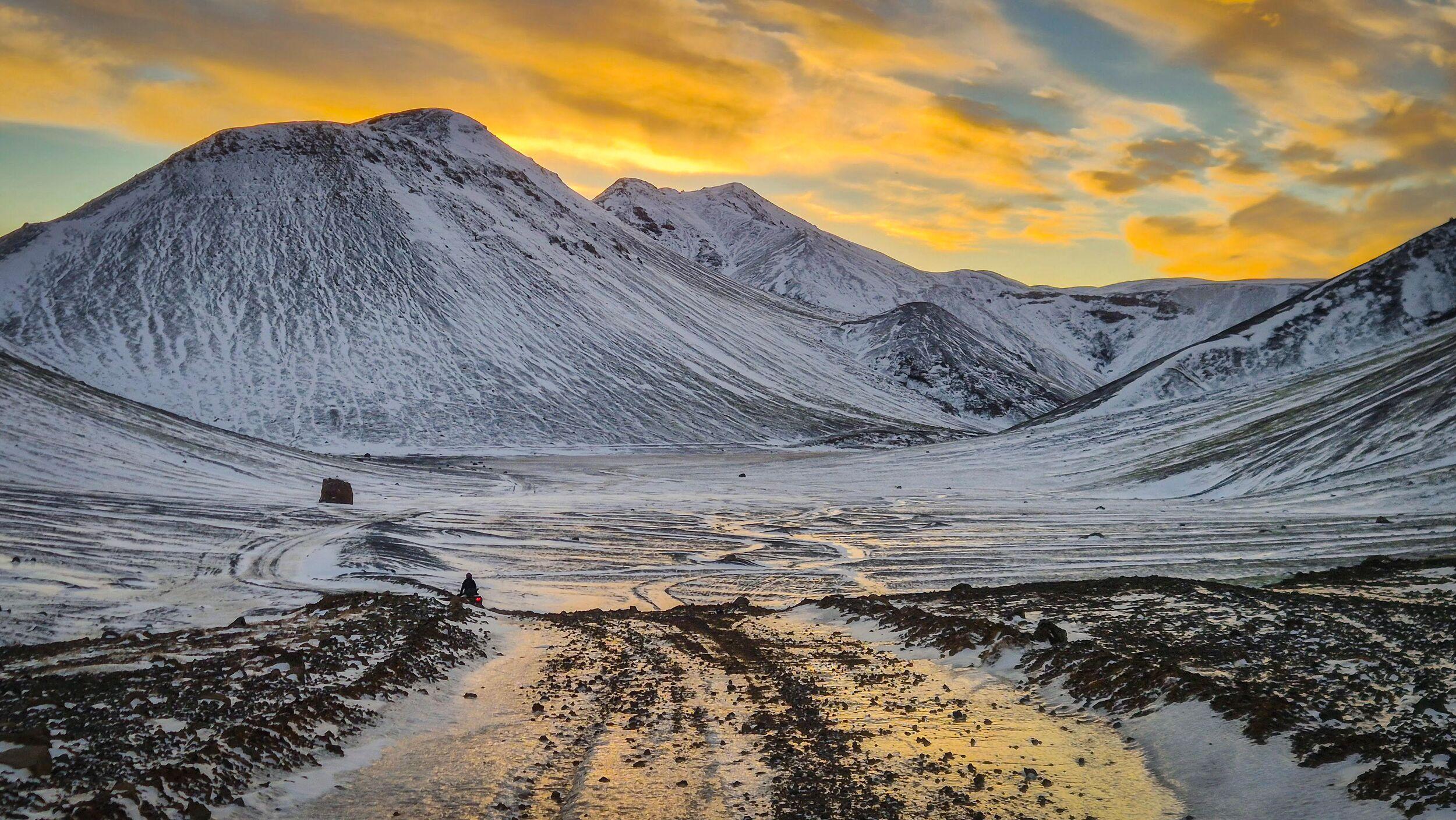 An icy gravel road winding through snowy mountains under a sky streaked with golden clouds