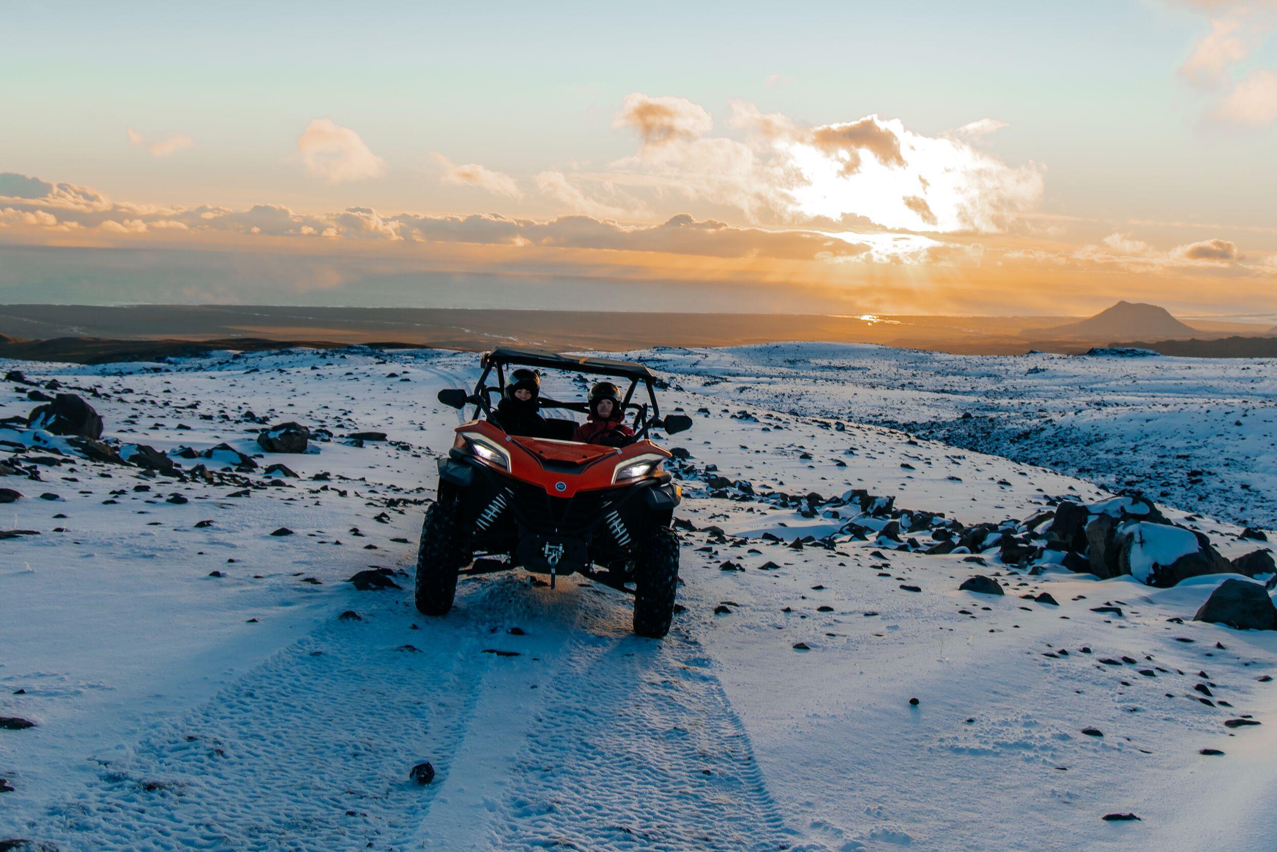 Two people in a buggy vehicle going over a snowy terrain with the sunset in the background