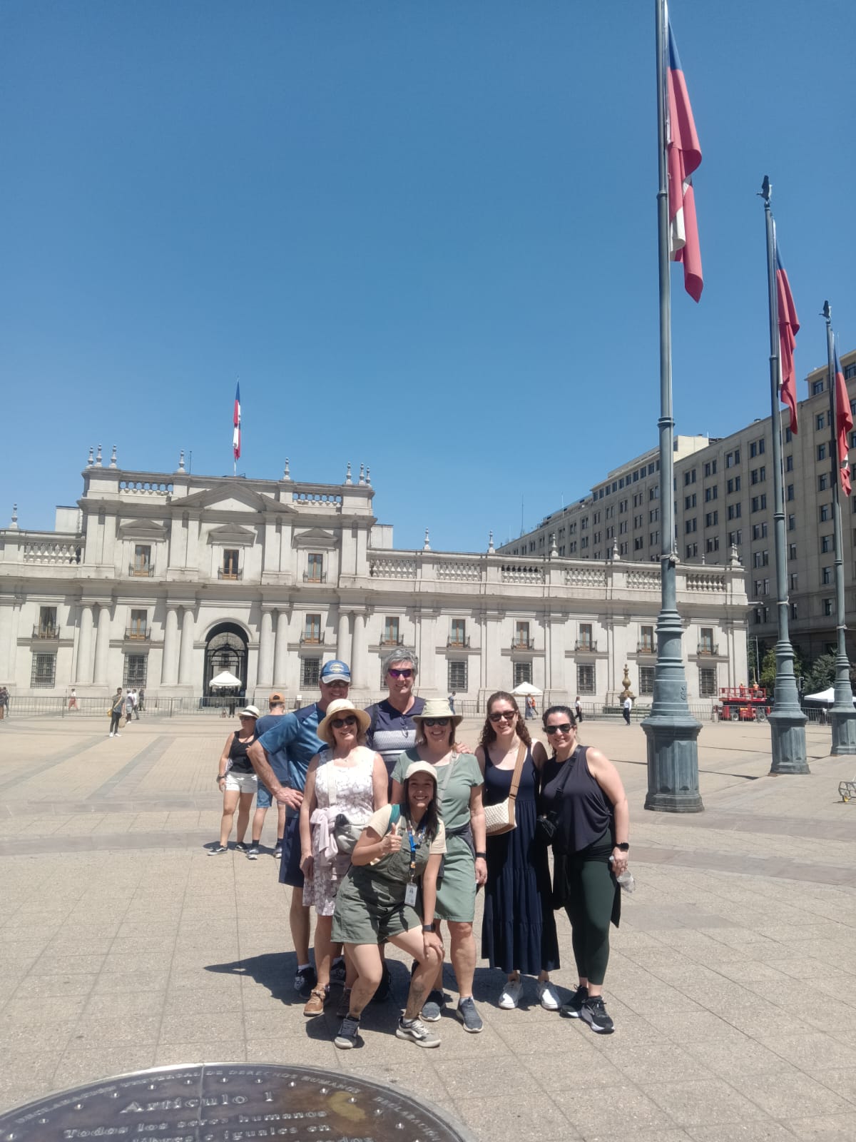 A group of travelers posing together in the plaza in front of La Moneda palace, Chilean flags flying above