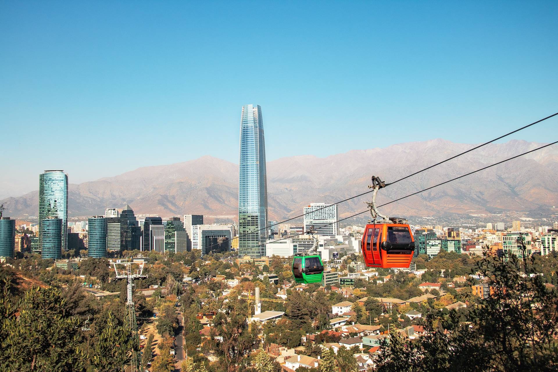 A red cable car crossing above the trees with the Costanera Center tower and the mountains behind it