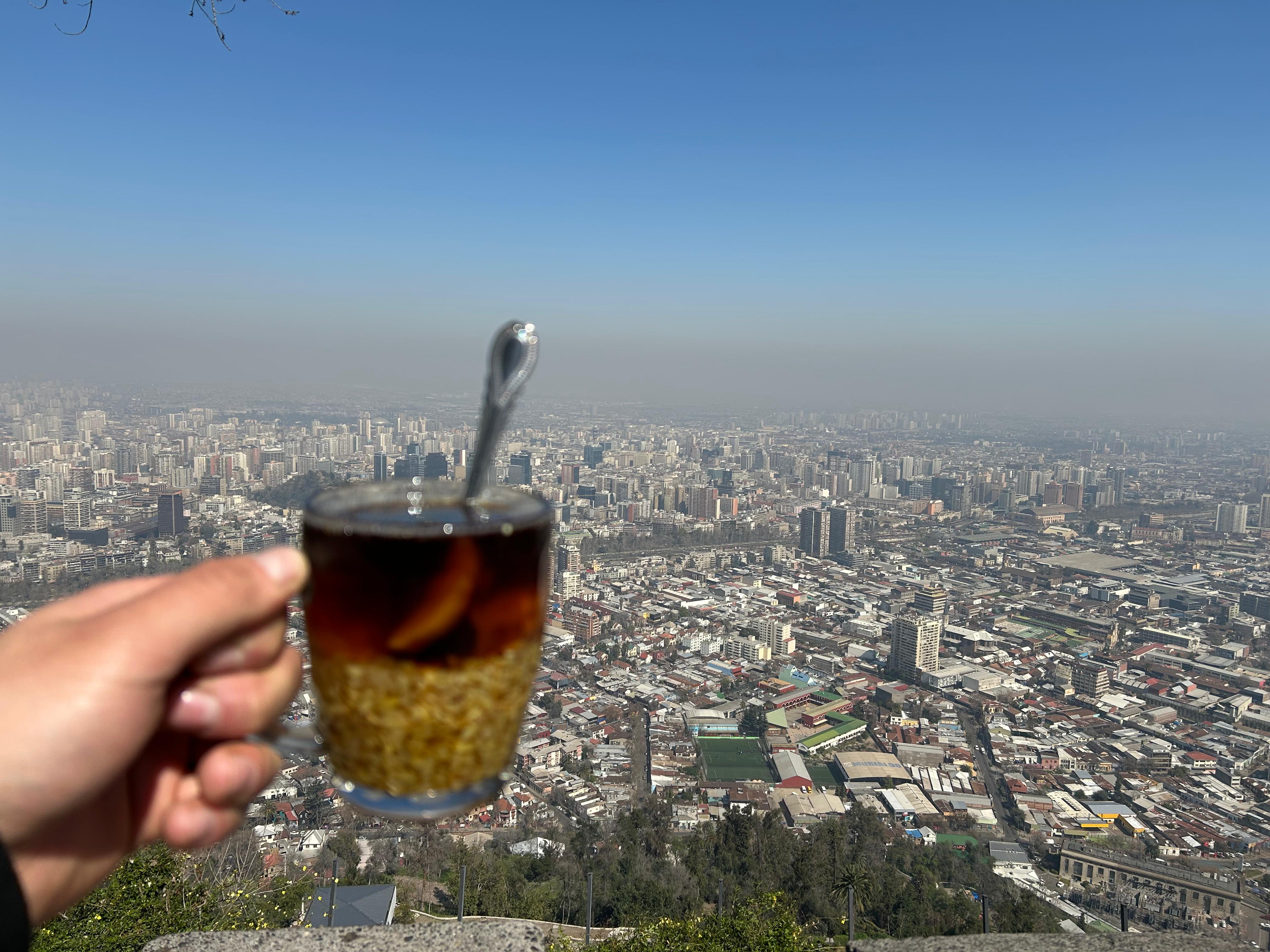 A hand holding a small glass of Chilean wine with the city spread out far below