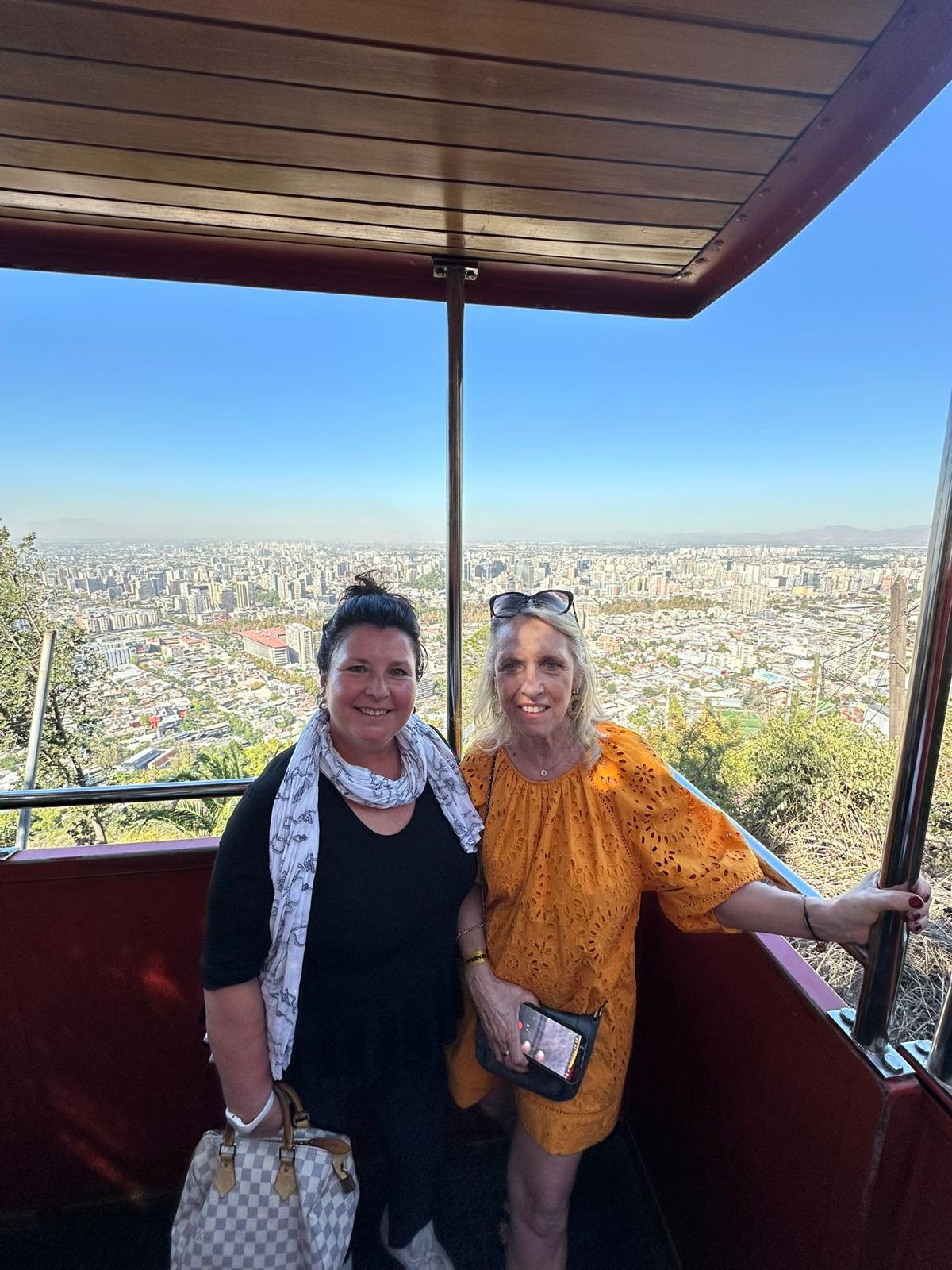 Two women posing together inside a cable car cabin with the city visible below