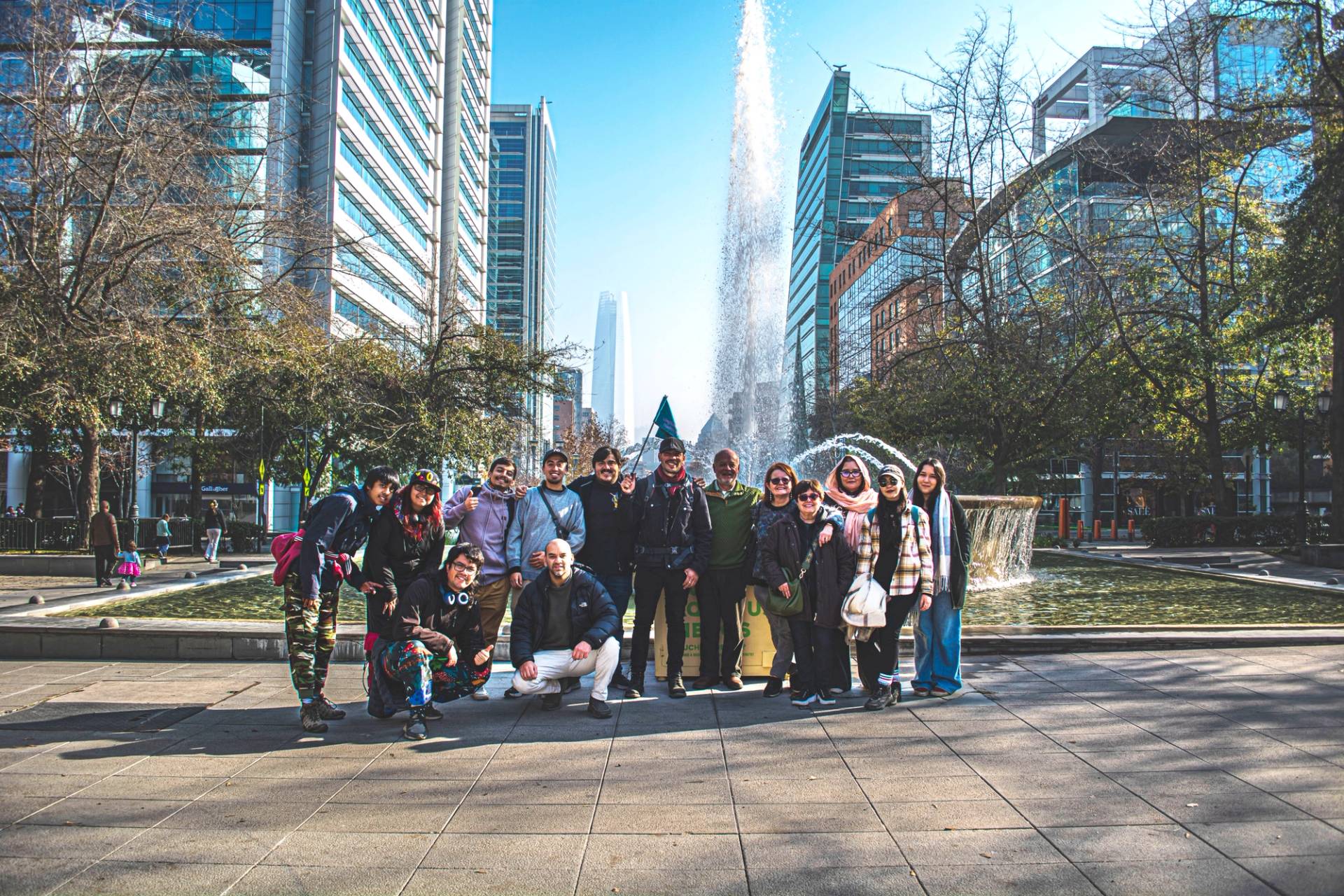 A tour group posing together in a plaza beside a fountain, glass office towers rising around them