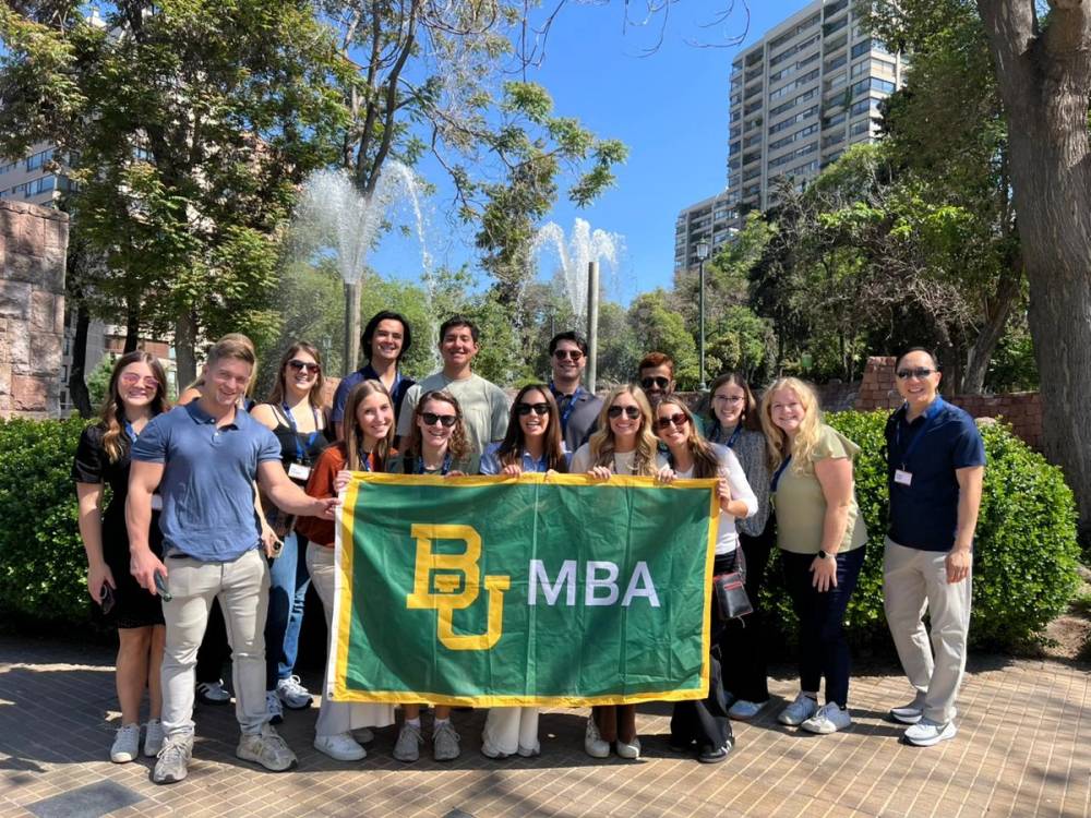 A university group holding their banner together in a Santiago park, a fountain behind them