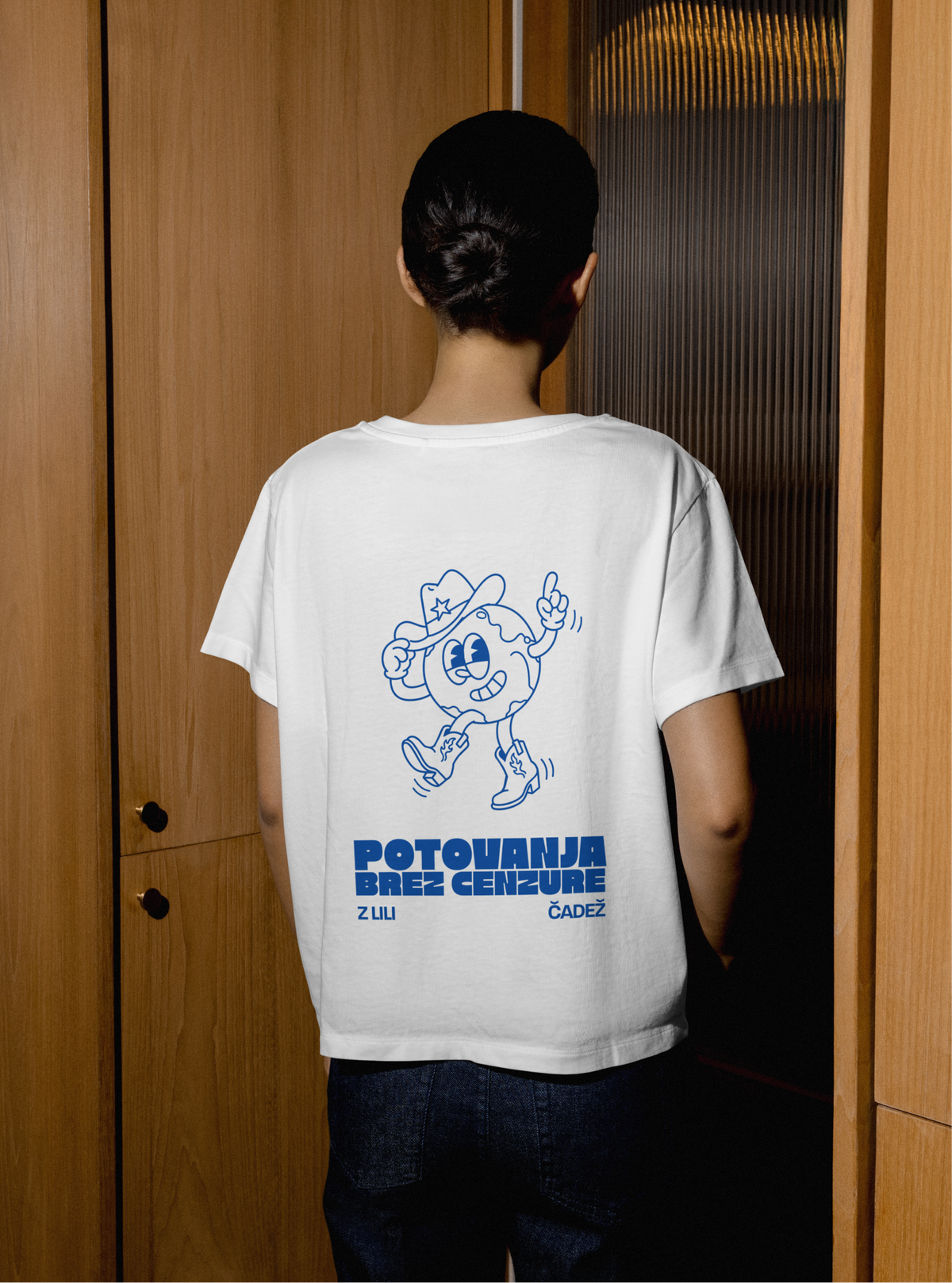 A woman seen from behind in a white T-shirt printed in blue with a globe in a cowboy hat and boots.
