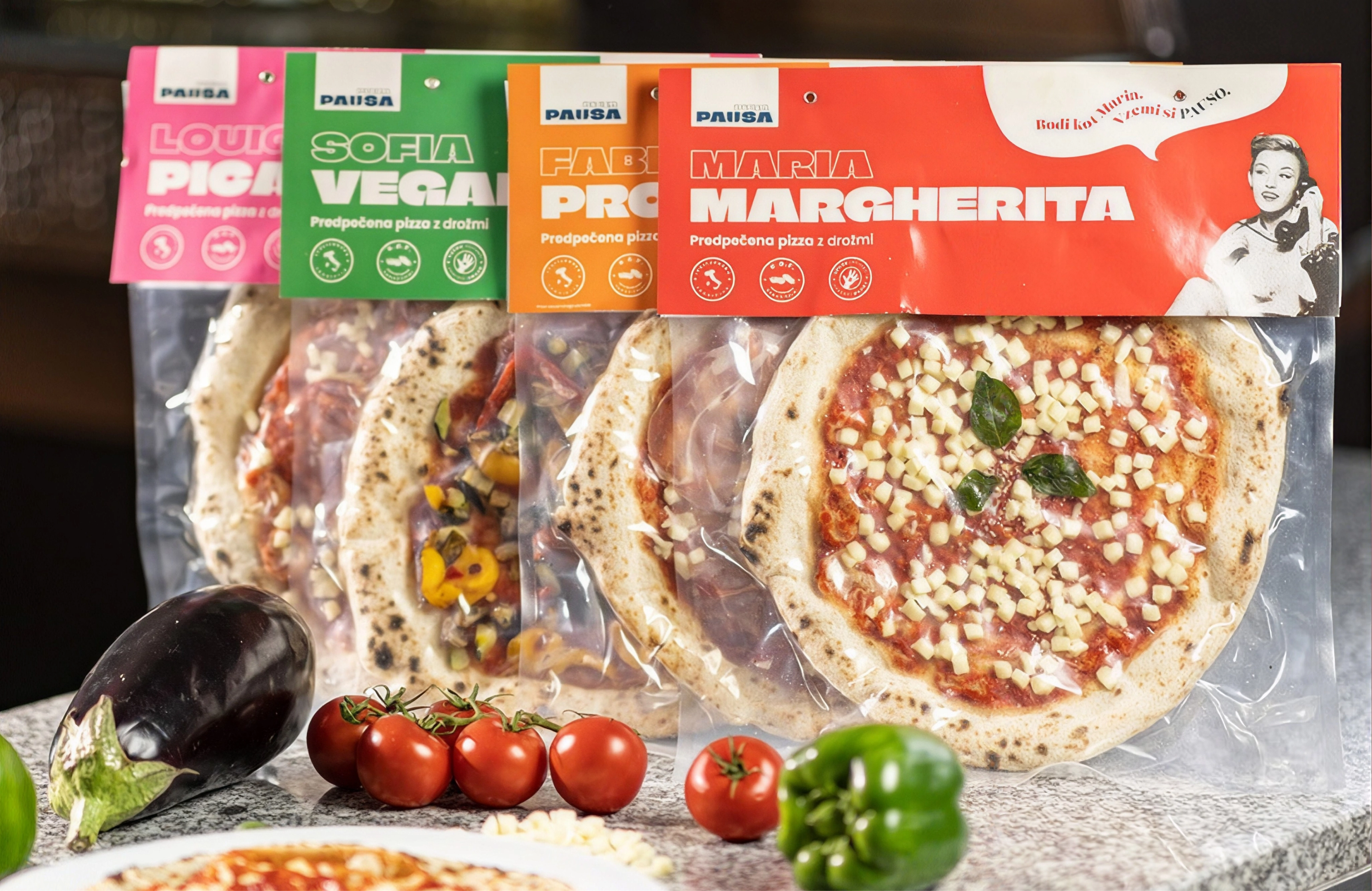 Pausa pizzas lined up on a granite counter, "Maria Margherita" in front, by tomatoes and an aubergine.