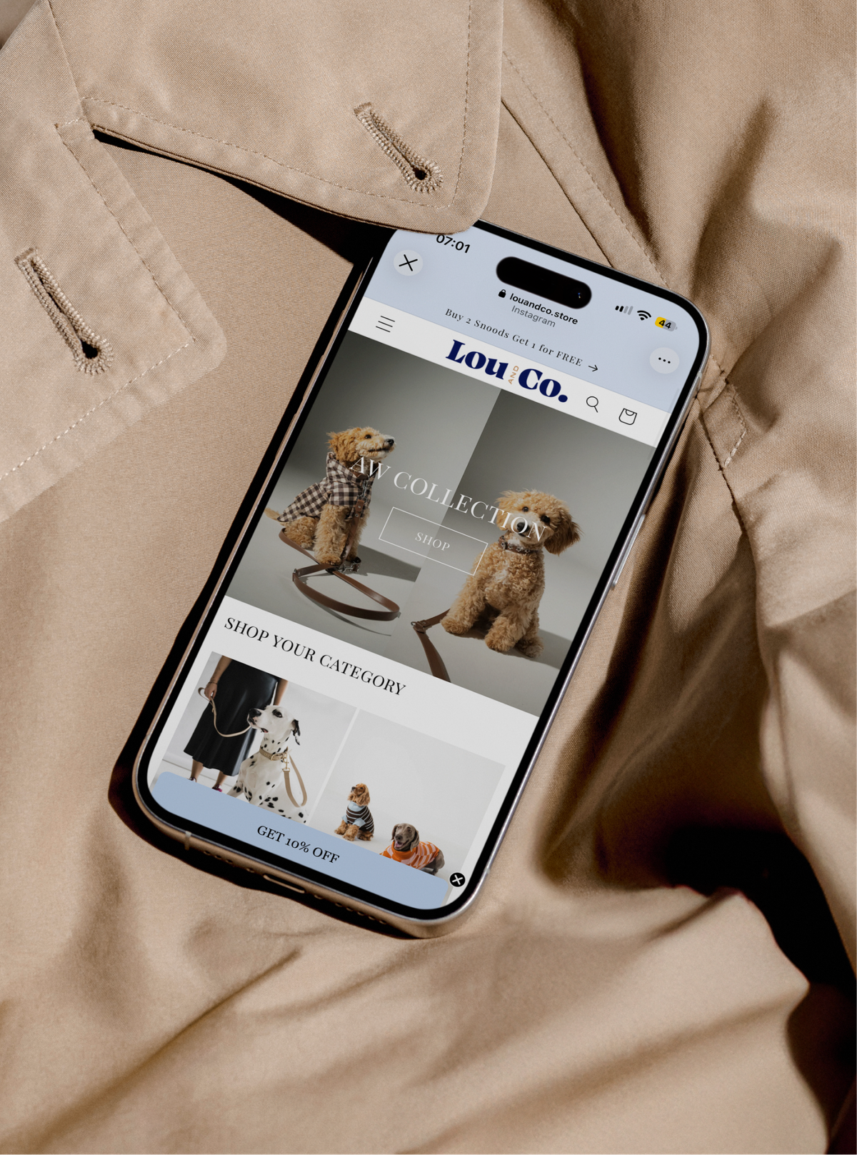 A phone on a beige trench coat showing the Lou & Co. online shop, with dogs in checked coats.