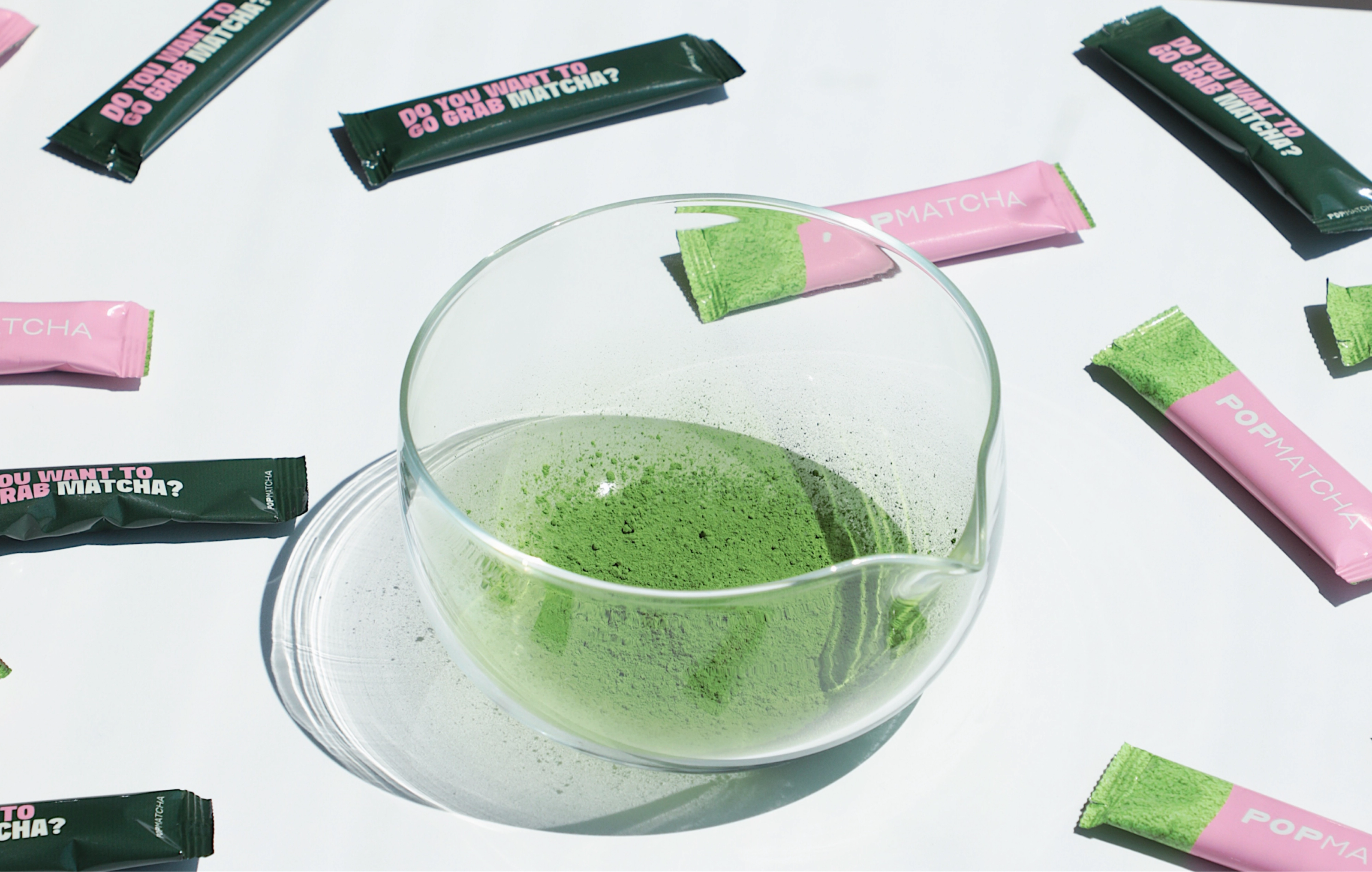 Pink and dark green Pop Matcha sachets scattered around a glass bowl of matcha powder on white.