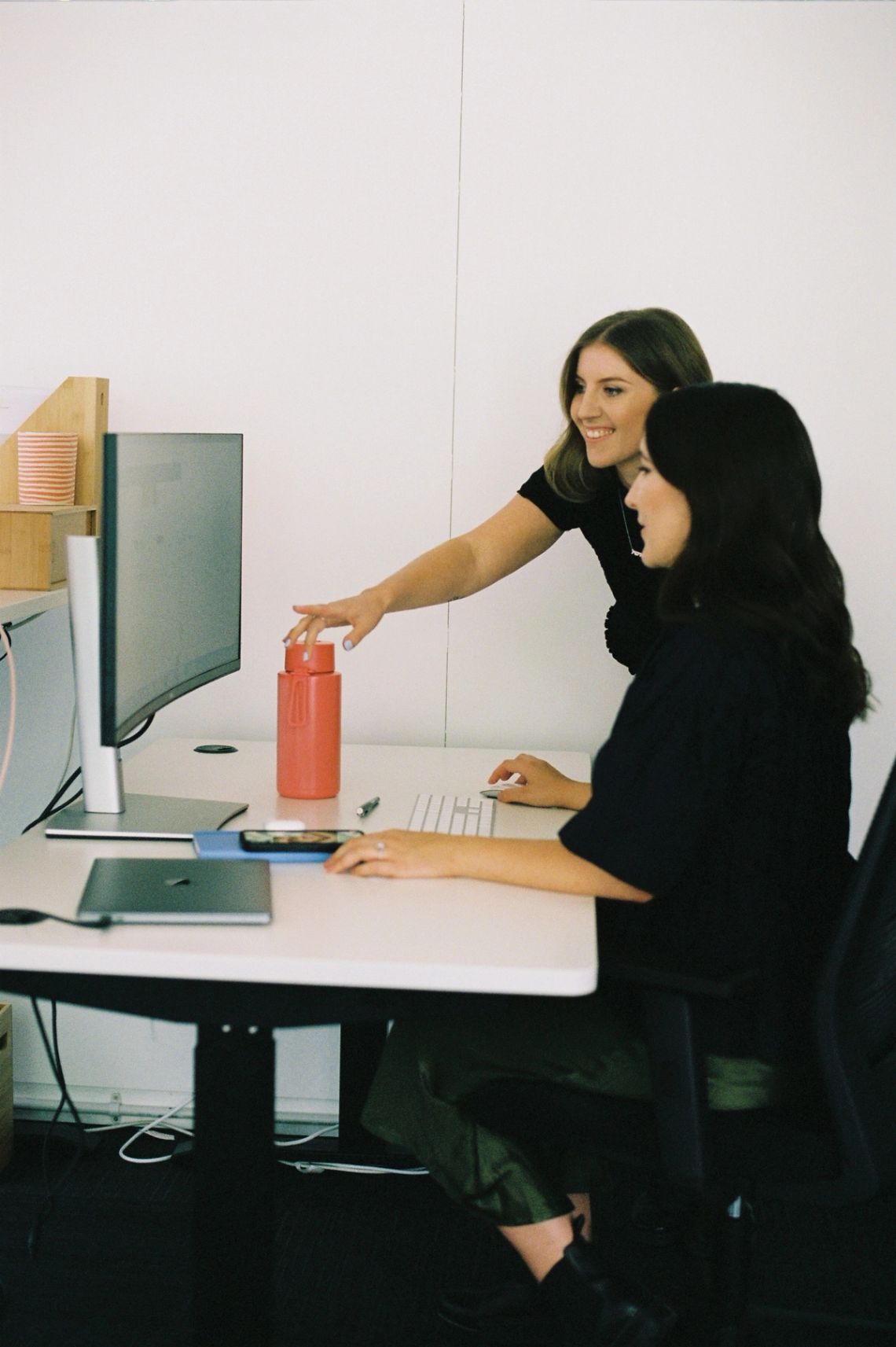 One person at a desktop workstation while a colleague leans in and points at the screen