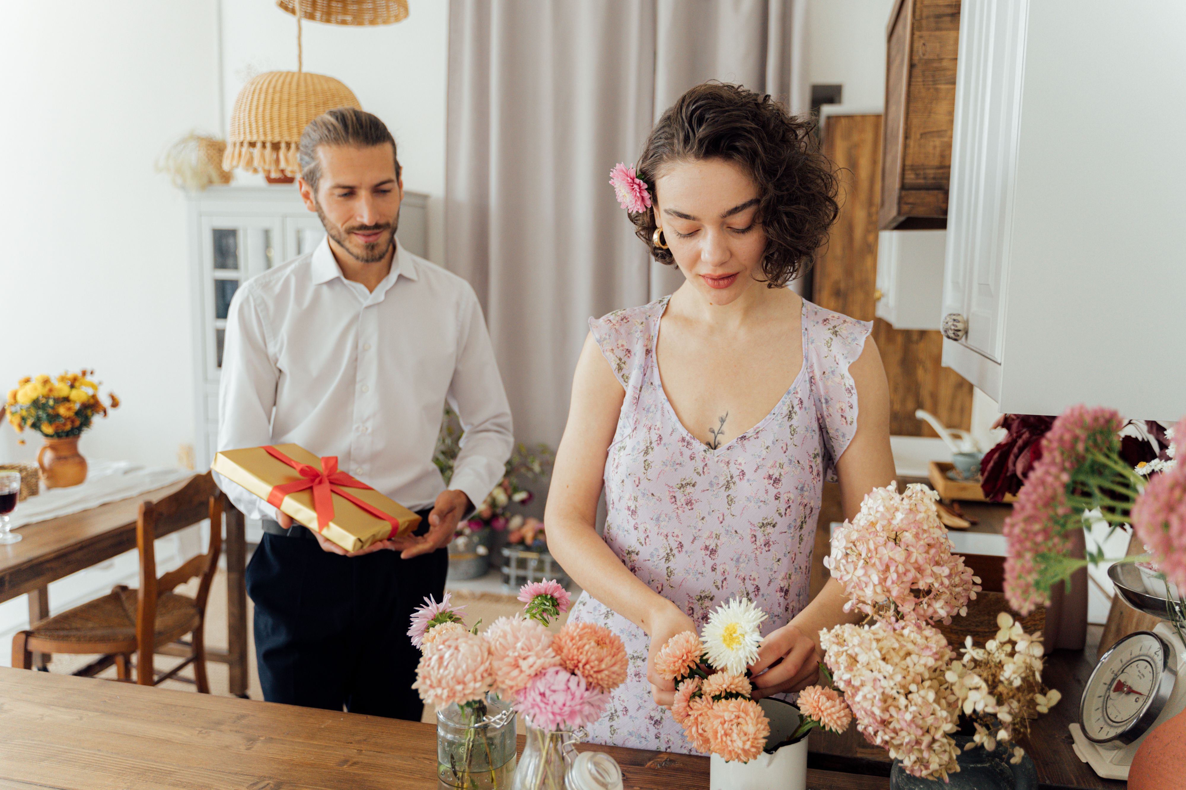 A man holding a wrapped gift while a woman arranges flowers in a bright kitchen