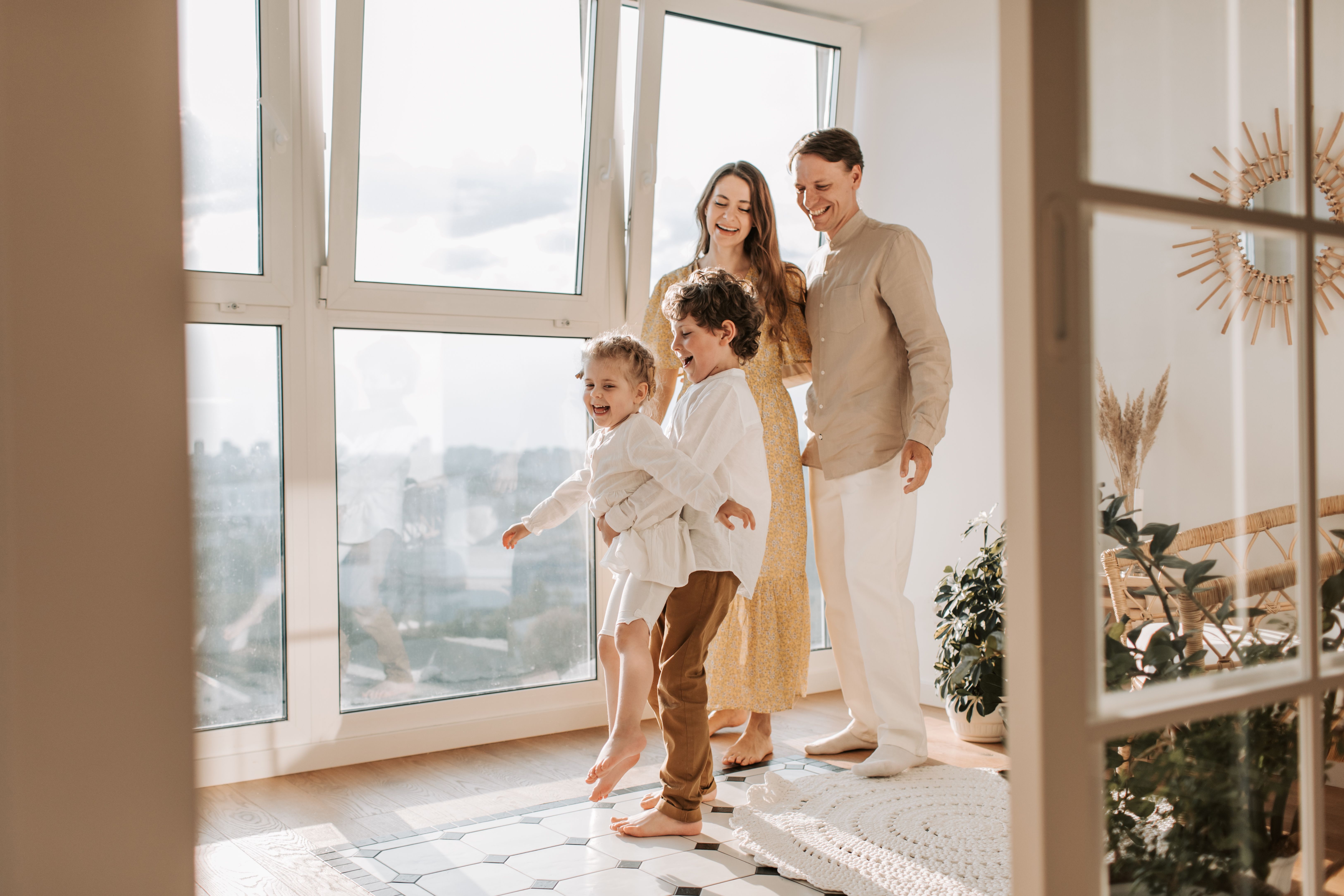 A family with two young children in a bright, tidy apartment by tall windows