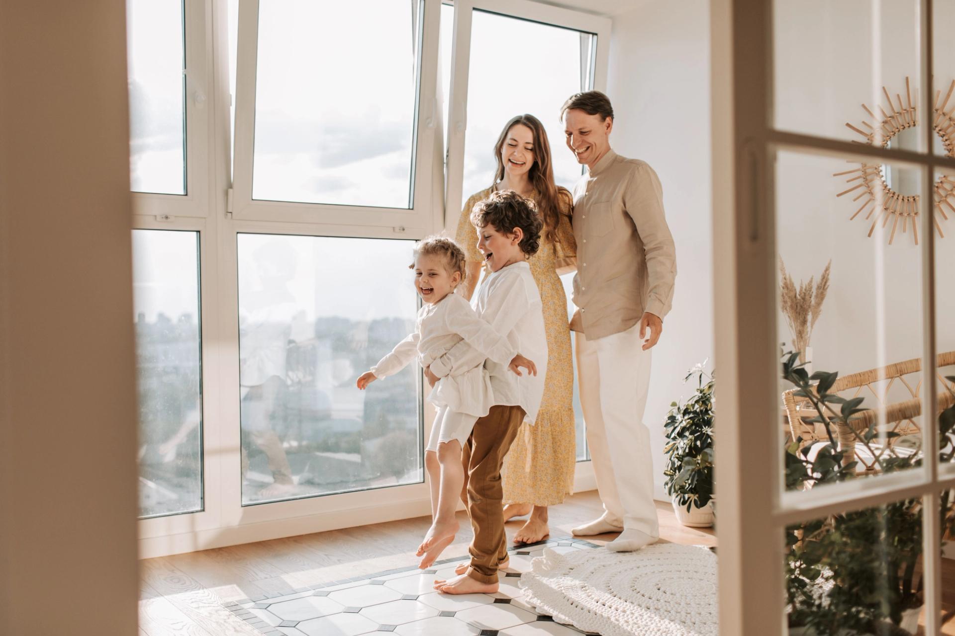 A family with two young children in a bright, tidy apartment by tall windows