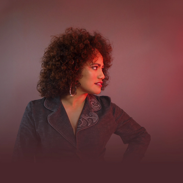 A woman with voluminous curly hair and red lips, wearing a dark jacket, gazes to the right with red light illuminating her face.