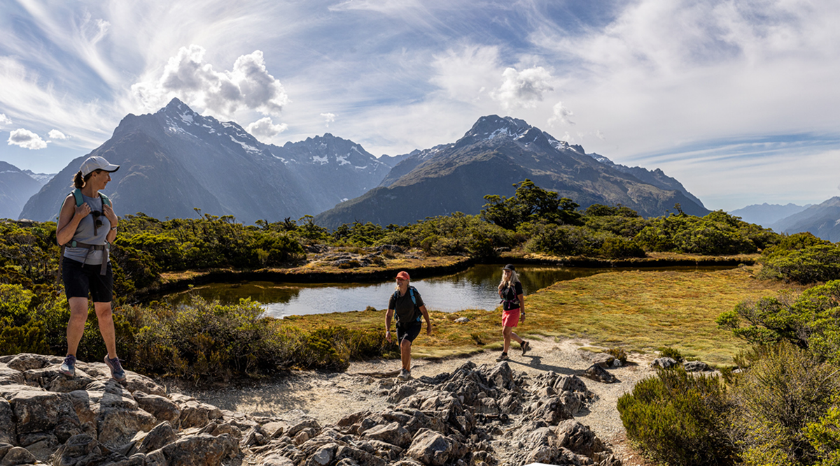 Three hikers on a trail through a mountainous landscape with a pond and snow-capped peaks.