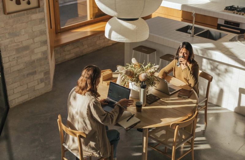 Two women working on laptops at a table in a bright, modern kitchen.