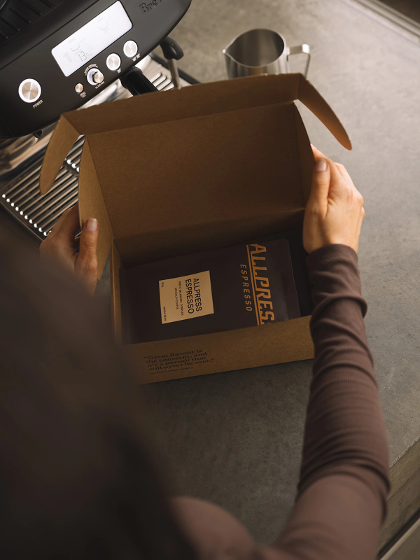 Hands opening a box of Allpress Espresso coffee next to a coffee machine.