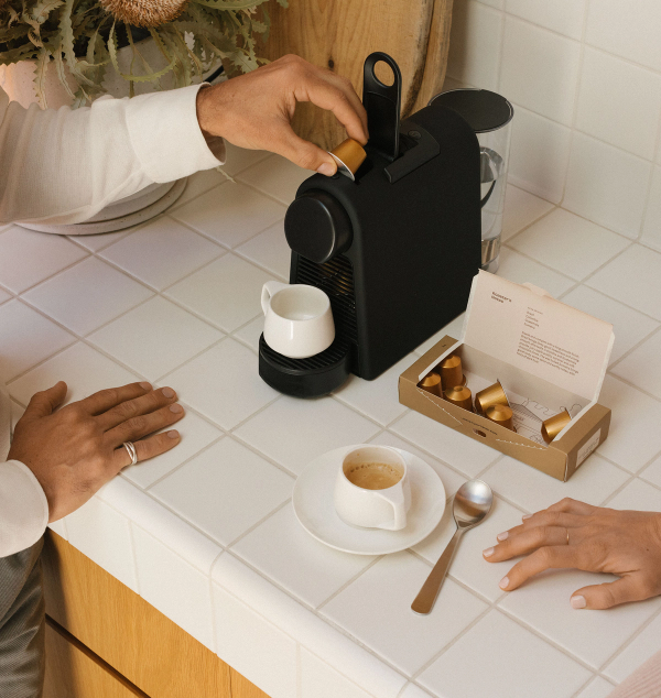 A hand places a coffee capsule into a black espresso machine on a white tiled counter, next to a cup of espresso and a box of capsules.