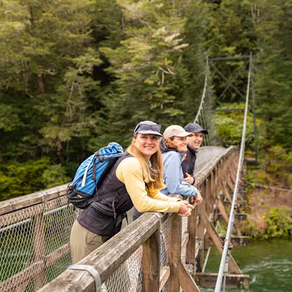 Three smiling hikers standing on a wooden suspension bridge over a river with a forest in the background.