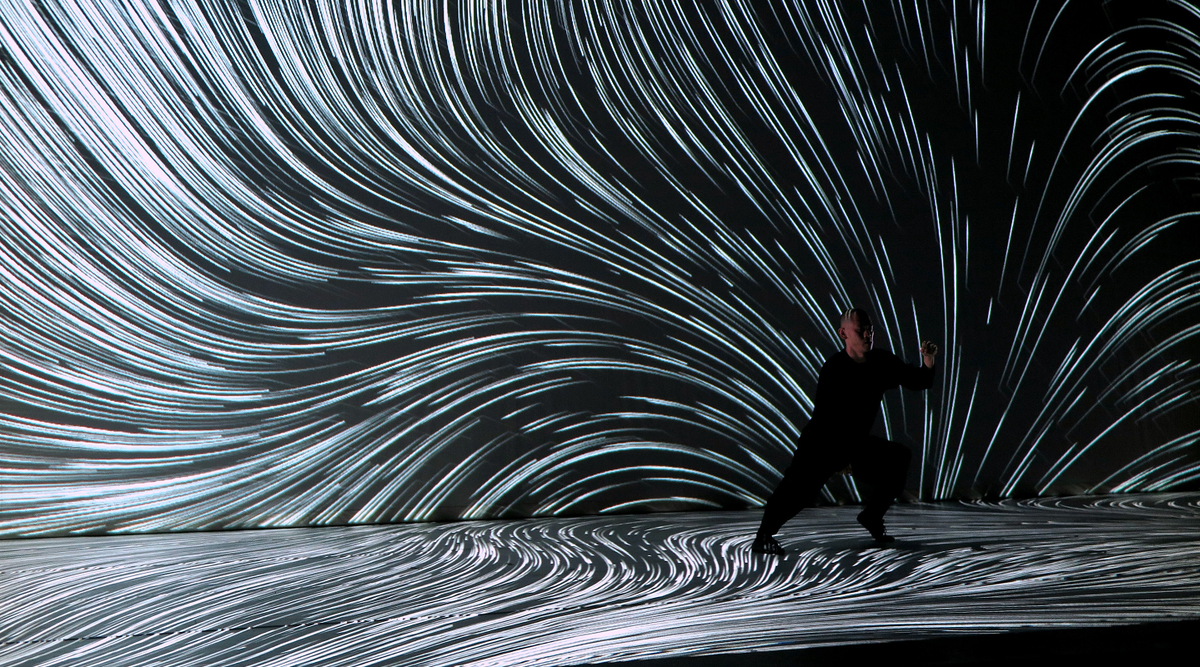 A person in black on a dark stage with swirling white light trails projected onto the wall and floor.