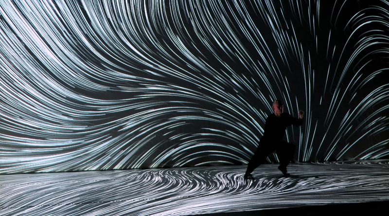 A person in black in a dynamic pose on a dark stage with swirling white light streams projected across the floor and wall.