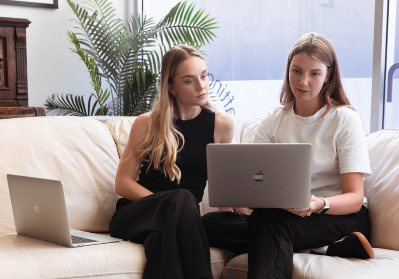 Two young women sitting on a couch with laptops.