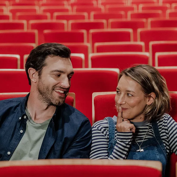 Man smiles at a woman holding a finger to her lips in a red movie theater.