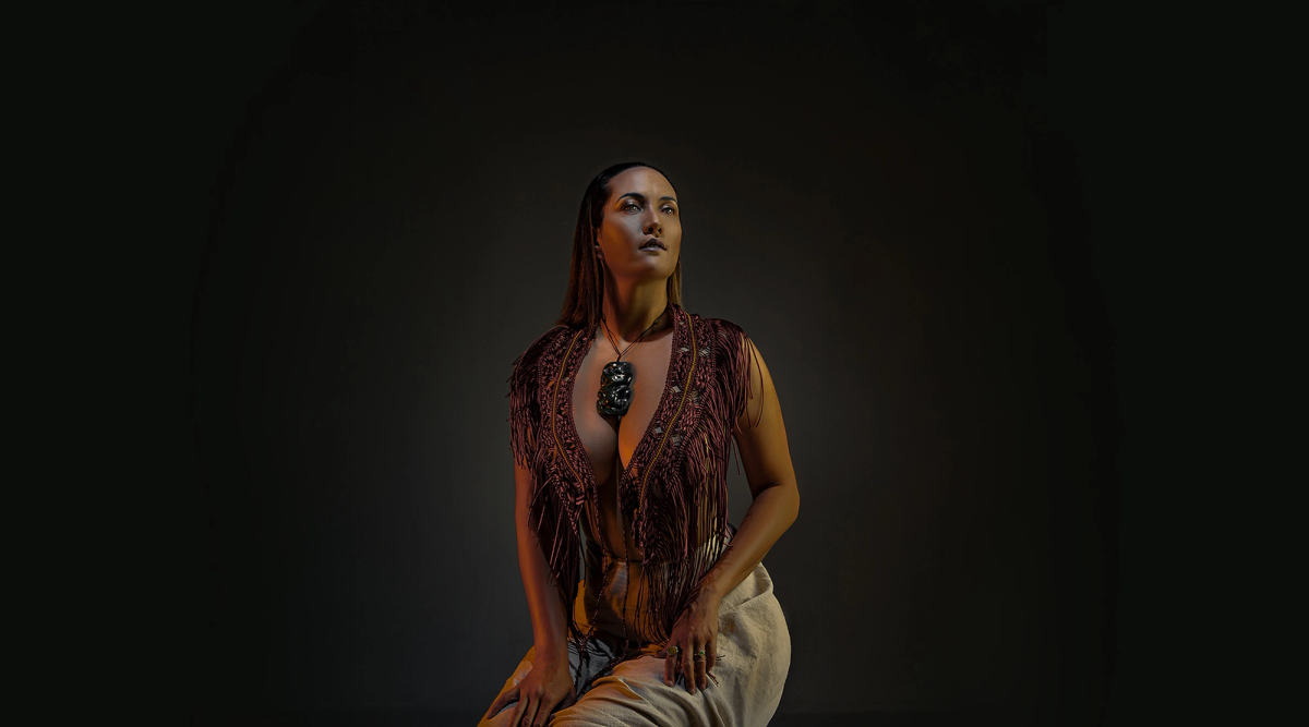 A woman with long hair, wearing a fringed top, light pants, and a dark pendant, kneels and looks up under warm orange lighting against a dark background.