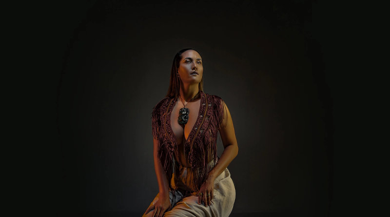 Woman in a fringed vest and ornate necklace kneeling, looking upwards against a dark background.
