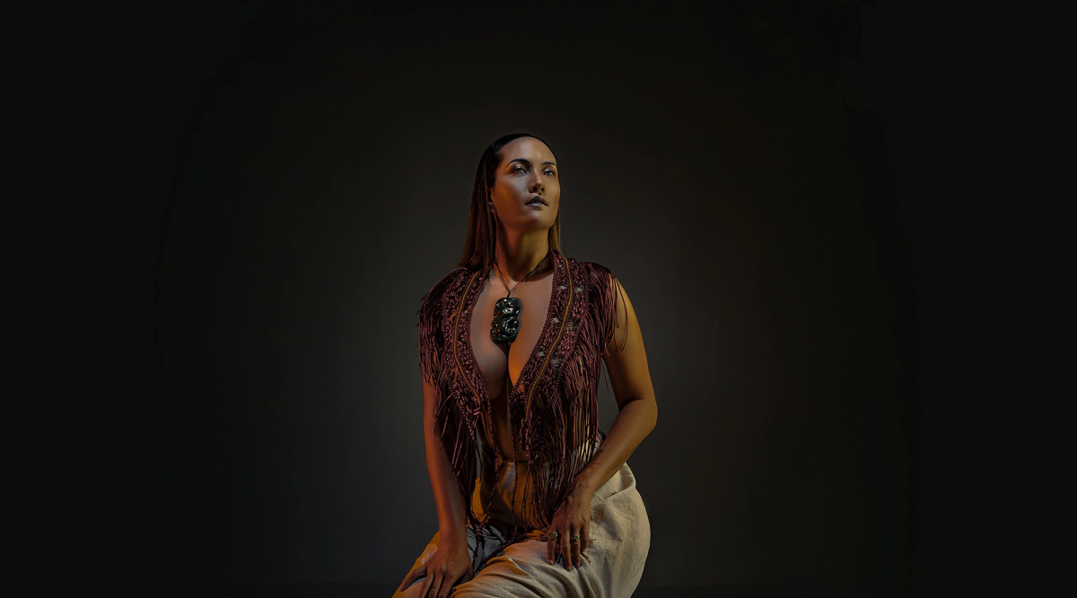 A woman with long hair, wearing a fringed top, light pants, and a dark pendant, kneels and looks up under warm orange lighting against a dark background.
