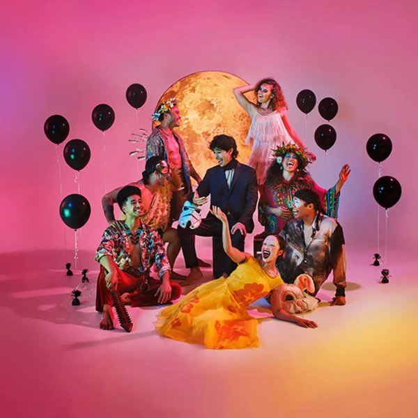 Eight people in diverse, festive costumes pose happily in front of a glowing moon and black balloons.