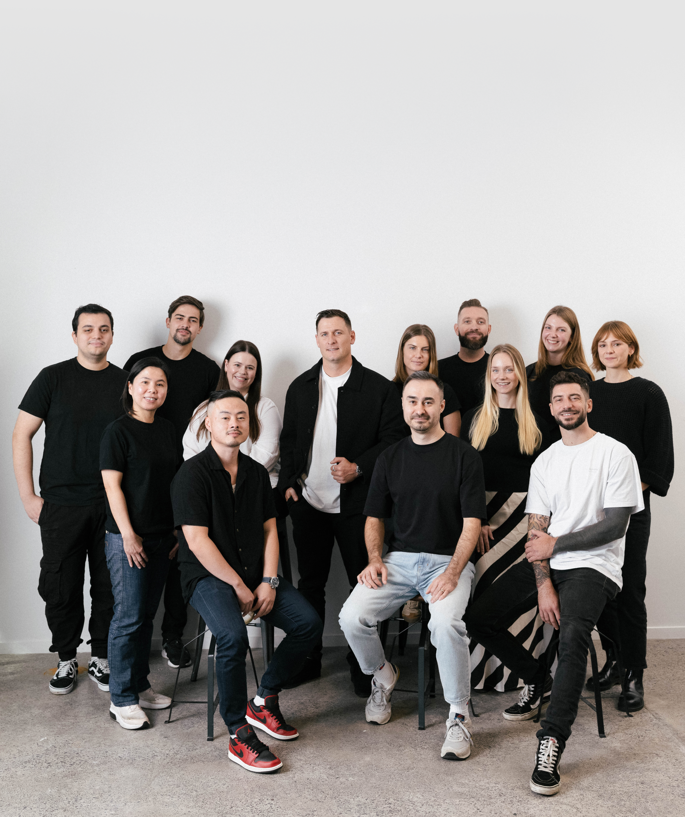 A diverse group of 14 people, some seated and some standing, smiling in black and white clothing against a white wall.