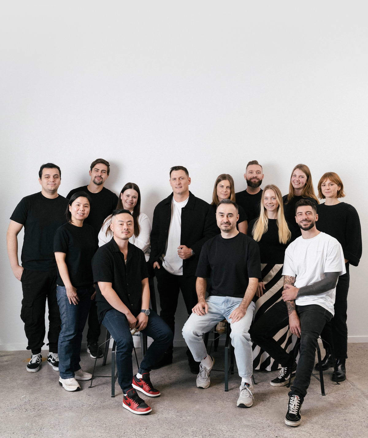 A diverse group of 14 people, some seated and some standing, smiling in black and white clothing against a white wall.