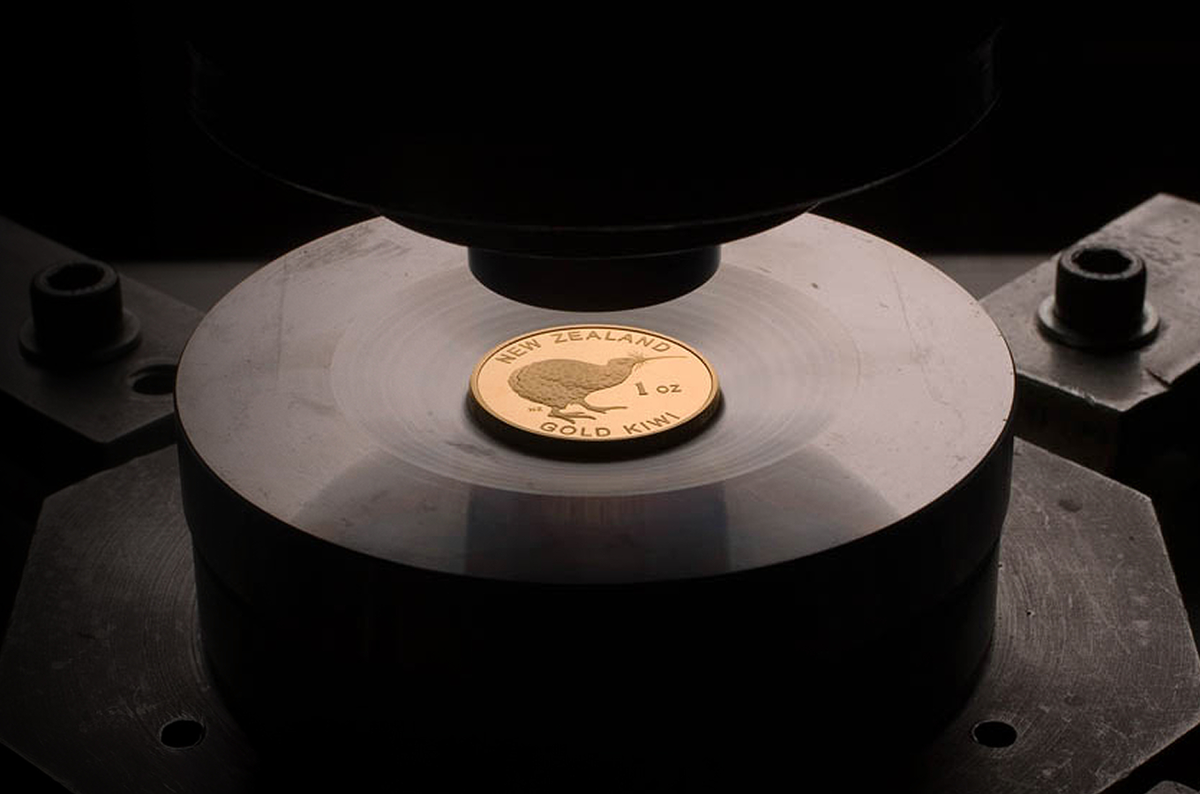 A New Zealand 1 oz Gold Kiwi coin with a kiwi bird design, resting on a metal platform under a minting press.