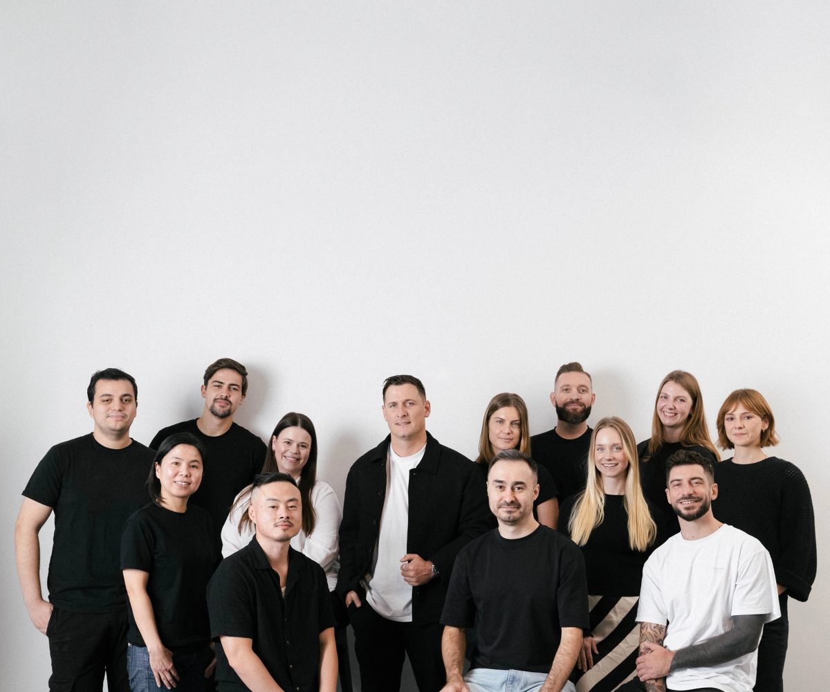 A diverse group of 14 people, some seated and some standing, smiling in black and white clothing against a white wall.