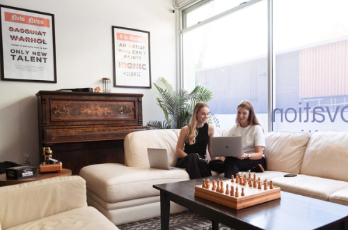 Two women on a cream sofa using laptops, with a chess set on the coffee table.