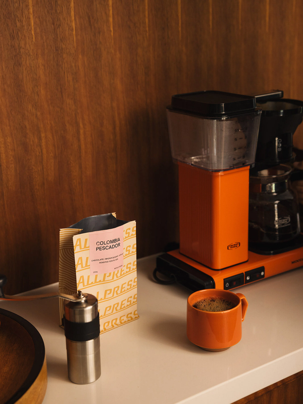 An orange coffee maker, a bag of Allpress Colombia Pescador coffee, a silver grinder, and a matching orange mug of coffee on a white countertop.