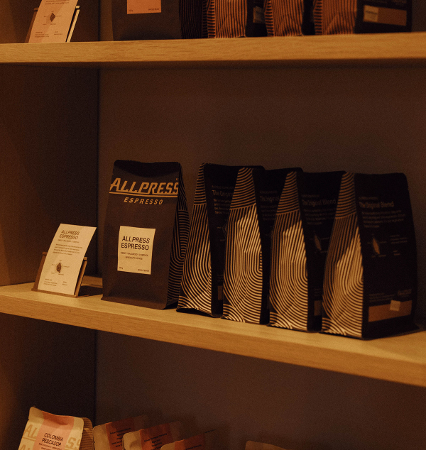 An orange coffee maker, a bag of Allpress Colombia Pescador coffee, a silver grinder, and a matching orange mug of coffee on a white countertop.