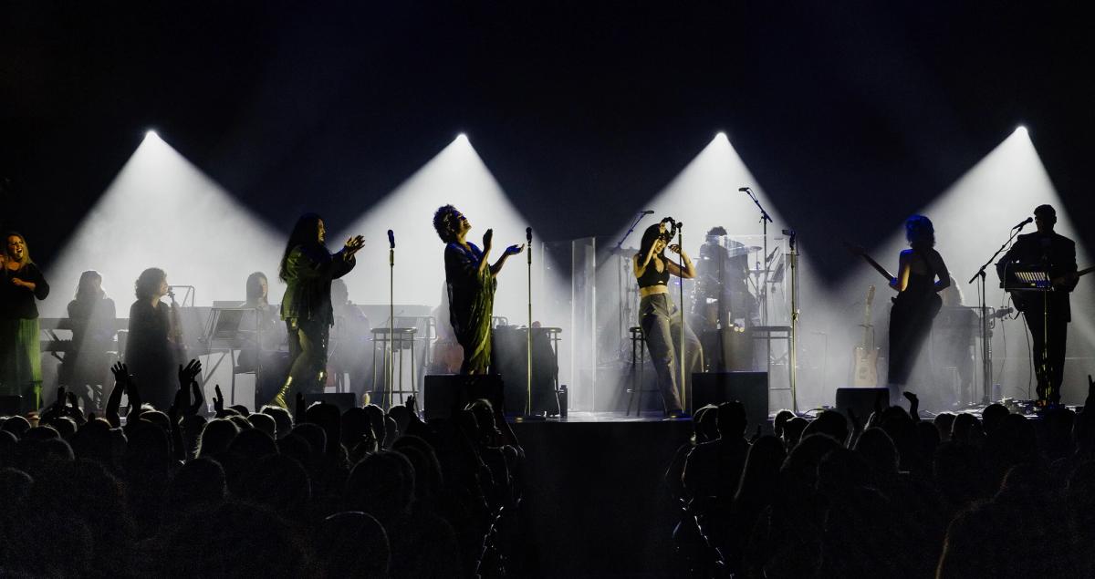 A live band and singers perform on stage under bright spotlights, with a silhouetted audience.