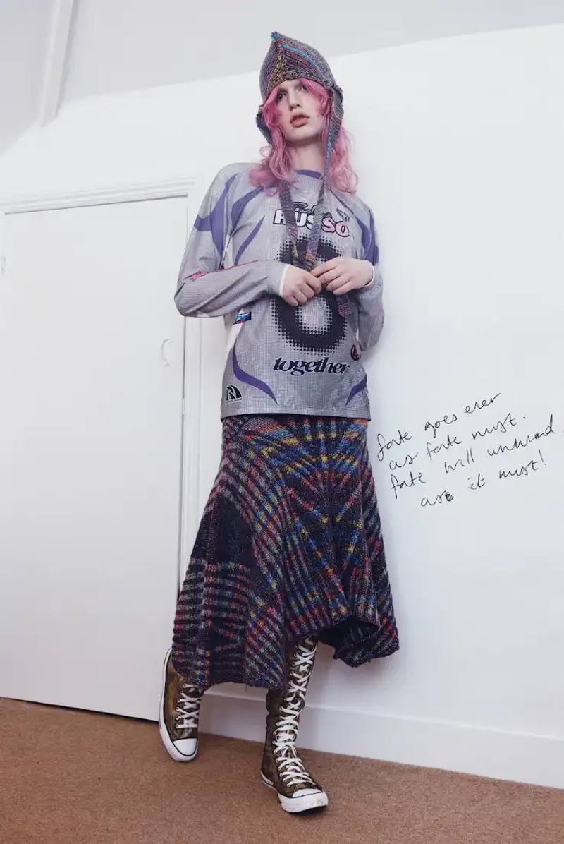 Photo of Paolina Russo's Autumn-Winter 2025 fashion collection, which combines medieval tailoring with early 2010s aesthetics. The collection features purple as a dominant color across various pieces, including denim, hoodies, and knits. Key elements include evolved "warrior knit" pieces, gold foil embellishments, and practical accessories like detachable hoods. The article also describes the designers' approach to merging traditional craftsmanship with modern technology, and mentions their recent industry recognitions including Forbes 30 under 30 and being finalists for prestigious fashion prizes.