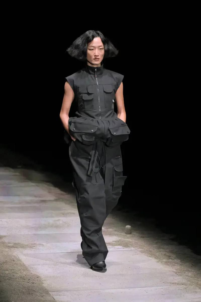 Models walk on sand-covered runway inside dark gymnasium, wearing layered winter clothing with misaligned buttons and multiple belts from Magliano's FW25 collection.