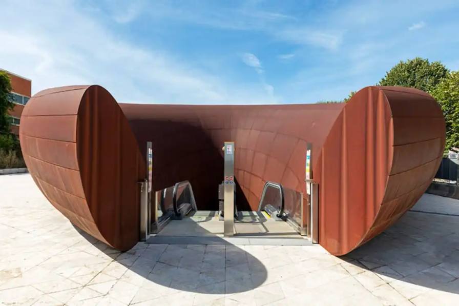 Two contrasting subway station entrances in Naples designed by Anish Kapoor: one features curved weathering steel panels with organic texture rising from plaza pavement, the other shows a sunken aluminum tube entrance with brushed metal finish reflecting surrounding buildings