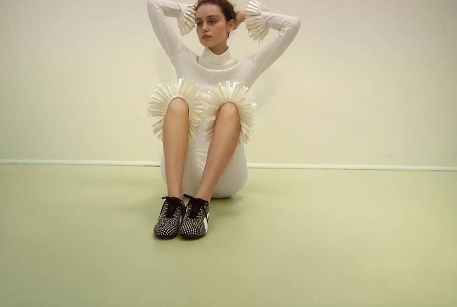 Photograph of model Alek Wek sitting on the floor wearing a white outfit with ruffled knee details and black and white patterned sneakers.