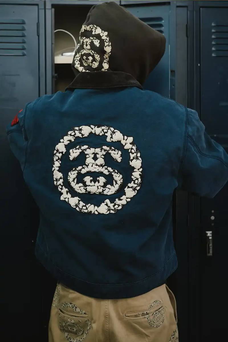 A display of Stüssy x Denim Tears' school-inspired collection, showing oxford shirts, blazers, and chinos arranged on a neutral background. The pieces feature co-branded designs and Pan-African American flag motifs.