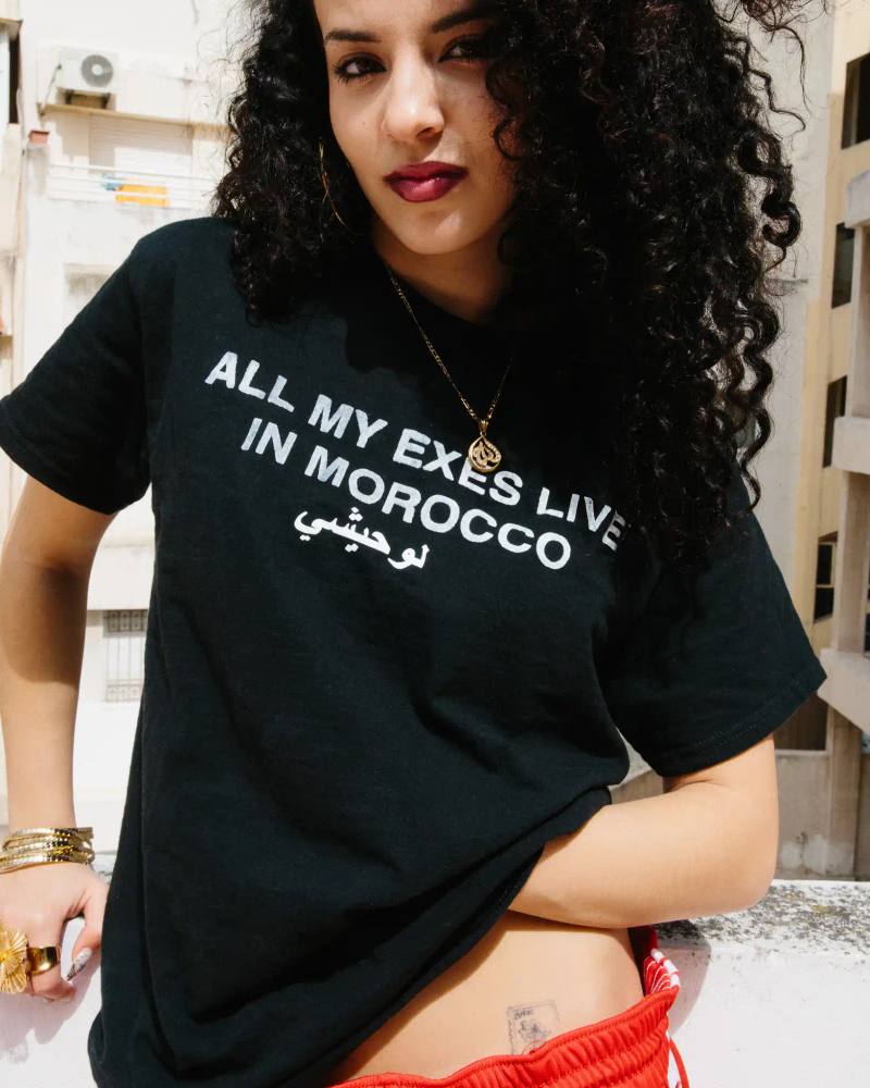 image of Zina Louhaichy, a multidisciplinary artist who blends her Moroccan heritage with New York City influences in her fashion designs, photography, and acting. The interview explores her experiences growing up between cultures, her creative inspirations, and her mission to represent North African diaspora stories. Zina discusses her fashion collections "All My Exes Live in Morocco" and "BLADI" that challenge Western stereotypes while celebrating her roots. She shares memories from Morocco, her creative process, and her commitment to creating authentic representation for MENA individuals in fashion and media.