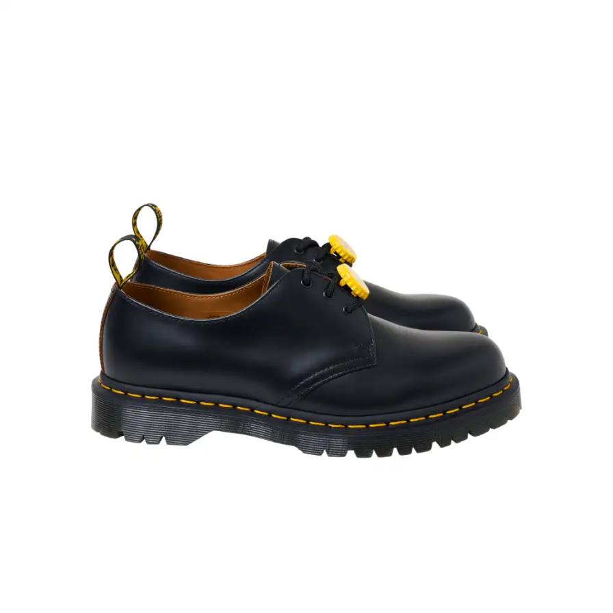 PALACE TEAMS UP WITH DR. MARTENS FOR FIRST-EVER COLLAB photo of the first collaborative collection between British brands Palace and Dr. Martens. The collection features three versions of Dr. Martens' classic 1461 shoe in black, cherry red, and woodland camouflage colorways. Each pair is made from vintage leather and includes distinctive bottle cap charms inspired by 1980s trends. The shoes feature a BEN sole with traditional yellow stitching and "Palace" text on the heel loop instead of the usual AirWair logo. The collection will be available online starting April 11 at 12:00
