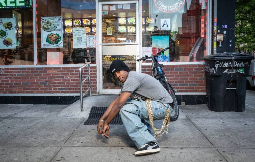 person squats on a New York sidewalk wearing baggy jeans, a grey shirt, a black beanie, and black and white Puma Suede sneakers. A bicycle and a shop entrance stand behind the person.