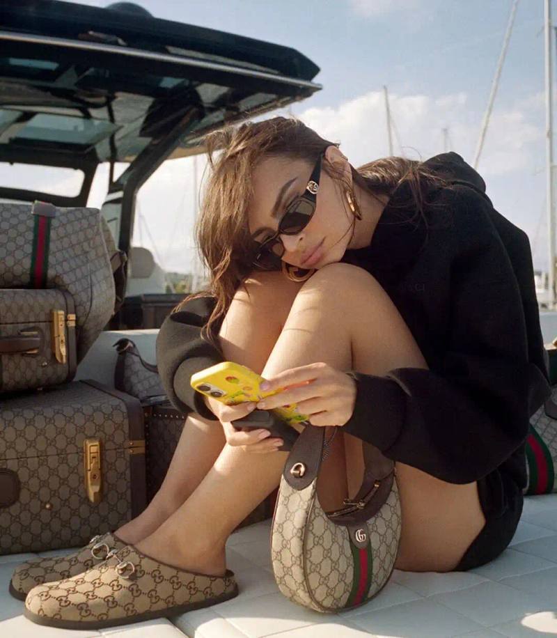 image of Emily Ratajkowski modeling Gucci handbags with the signature GG monogram pattern. Photographed in Cannes by Daniel Arnold, the campaign highlights heritage-inspired bag designs including the new Giglio model and updated Ophidia series. Images show the model in various city and coastal locations demonstrating how the monogram pattern adapts to different settings and occasions.